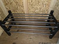 Photo of the IKEA TJUSIG shoe shelf showing black metal end supports and horizontal stainless steel rods, placed against plywood background.