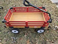 Red wooden wagon with black metal handle and removable red rails on natural wood base, shown on grass.