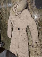 Full view of long off-white puffer jacket showing front pockets, belt, hood with fur trim, and overall quilted design.
