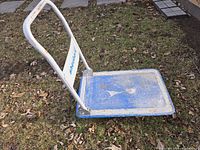 Side angled view of folding push cart hand truck showing white metal frame and blue platform resting on grass with signs of wear