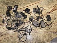 Overview of all 12-13 miscellaneous chargers and adapters spread on stone surface showing variety of shapes, cords, and sizes