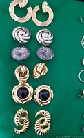 Six pairs of assorted earrings laid out on a green surface showing different designs and metal finishes.