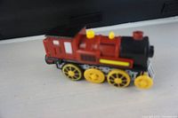 Side view of red cast metal toy train engine with yellow wheels on surface, showing yellow chimney and external details