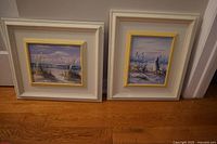 Pair of paintings side by side on floor showing white wide wood frames with yellow inner frames and pastel coastal scenes