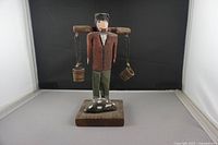 Front view of Emile Bluteau wooden folk art sculpture of man carrying water buckets on shoulder pole, mounted on wooden base with carved stone object.