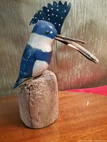 Side view of hand carved and painted King Fisher bird mounted on rustic wood base, showing blue and white color pattern with dot detail on crest, holding a painted fish in beak.