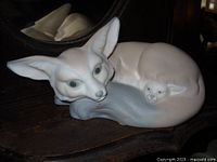 Side view of the LLADRO mother fox and cub figurine showing the cub nestled under the mother's tail on a wooden surface with reflection in the background.