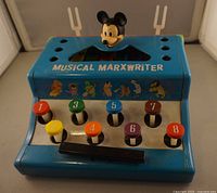 Front view of blue Musical Marxwriter toy featuring colorful numbered keys and Mickey Mouse head topper, with visible detail on illustrations and keys.