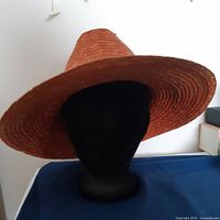 Front view of orange 100% straw hat showing wide brim and pointed crown on black mannequin head against white background.