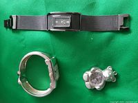 Photo of two rectangular wristwatches with silver mesh band and a small teddy bear pendant watch on green fabric background.