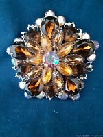 Front-view photo showing the brooch's detailed arrangement of amber-colored rhinestones with a central iridescent stone displayed on blue fabric.