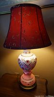 Full view of the lamp showing cranberry glass base with white porcelain overlay and the burgundy embroidered shade illuminated.