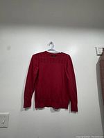 Full view of red sweater hanging on plastic hanger against white wall.