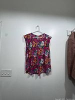Front view of a sleeveless floral blouse in rose pink color hanging on a white plastic hanger against a white wall.