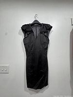 Black sleeveless French Connection dress hanging on black hanger against white wall. Features structured ruffles on shoulders, V neckline, fitted waist and knee-length hem.
