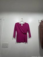 Front view of a bright pink blouse with a wrap style and decorative ring at the waist hanging on a white hanger against a white wall.
