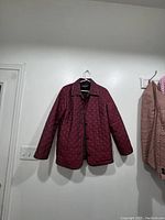 Full view of burgundy quilted coat hanging on hanger against white wall showing overall condition and shape.