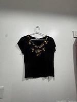 Front view of black short-sleeve blouse hanging on plastic hanger against white wall. Embellished neckline with metallic studs visible.