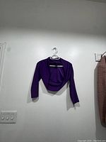 Full view of purple crop cardigan hanging on a hook against white wall.