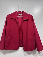 Front view of red women's jacket hanging on a hanger against a wall showing collar, sleeves, and front pockets.