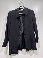 Front view of a black blazer hanging on a white hanger against a white wall, showing the button-up style and collar.