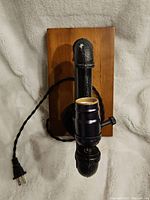 View of the lamp showing black metal pipe construction and wooden mounting base with attached electrical cord and plug.