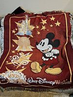 Photo showing large burgundy Walt Disney World tapestry throw featuring Mickey Mouse and iconic park landmarks with fringed edges.