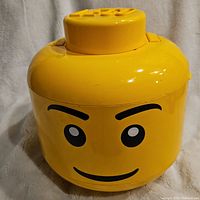 Front view of yellow Lego head-shaped container with printed face and Lego stud top.