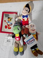 Plush toys of Pinocchio, Jiminy Cricket, and Geppetto, arranged on a white surface with visible Disney tags.