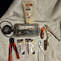 Overview photo of the lot's tools and miscellaneous items on a light background, showing the wire crimping tool, headlamp, Xacto blades, cable ties, locking pliers, hand knife, leather strap, and clear tool kit case.