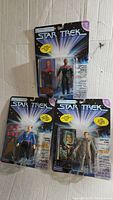 Four vintage Star Trek action figures new in package including Captain Picard (two versions), Captain Kirk, and Harry Mudd, arranged on cardboard backing with space background