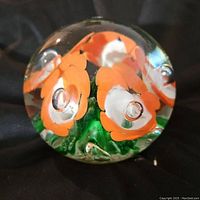 Close-up of front side of rounded glass paperweight showing embedded orange and white flowers with green stems and bubbles inside the glass, no damage visible.