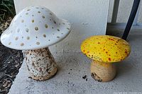 Two garden mushroom statues placed outdoors on a concrete surface, showing relative size and condition.