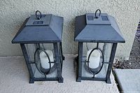 Pair of black metal outdoor lanterns with clear oval front design and candle-shaped inside light, showing full front view against a beige wall.