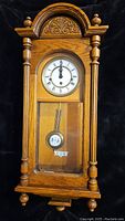 Front view of wooden wall clock showing carved top, dial with Roman numerals, and pendulum behind glass.