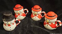 Four ceramic snowman mugs on black background, showing details of hats, scarves, and candy cane handles.