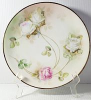 Full front view of round charger plate with white and pink roses and green leaves, with gold trimmed edge, on transparent stand