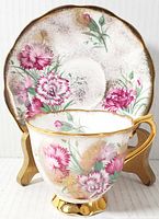 Photo showing front view of tea cup in front of saucer with pink carnations floral pattern and gold trim.