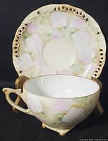 Tea cup placed in front of saucer, showing white embossed roses and pastel green and pink floral background with gold rim on saucer and cup rim.