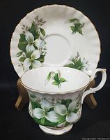 Front view of tea cup and saucer showing white trillium floral pattern with gold rim accents on black background.
