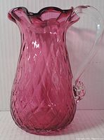 Front view of the deep cranberry-colored glass pitcher displaying the textured diamond pattern and scalloped rim, sitting on a plain background.