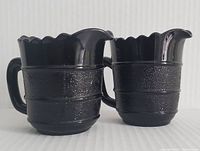 Front and side view of two matching black amethyst glass pitchers with scalloped rims and textured surface bands.