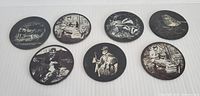 Seven round slate coasters with various engraved Celtic and historic scenes, arranged on white fabric.