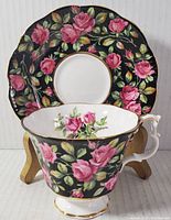 Front view of tea cup and saucer showing pink rose floral pattern and gold trim on black background.