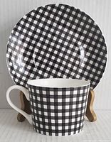 Black and white gingham check cup in front of matching saucer on small stand, showing front side of cup and full saucer