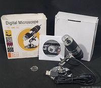 Digital microscope with LED lights on an adjustable metal stand, connected via USB cable, accompanied by software CD and original packaging box.