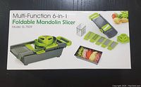 Box front showing the foldable mandolin slicer in green and gray with interchangeable blades and a container for collecting sliced vegetables.