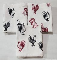 Three white cotton tea towels laid flat, showing red and black rooster prints across each towel.