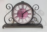 Full front view of retro metal mantle clock on flat surface, showing circular face with Roman numerals and curled metal side details.
