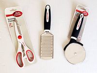 Top-down view of three kitchen tools: scissors, grater, and pizza cutter, all Betty Crocker brand, in packaging except the grater.